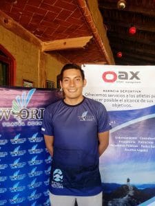 En esta imagen, se muestra a un hombre sonriente de pie frente a un banner de "World Travel Agency" y "Oax Sport Management". Lleva una camiseta azul con el logo de "World Travel Agency" y "Pescado de Moctezuma". Está de pie en un ambiente que parece ser una sala interior con paredes de ladrillo y techo de madera.