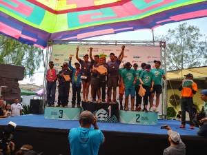 En esta imagen, se observa a varios equipos en el podio de una competencia. Hay tres niveles en el podio, numerados del 1 al 3, y los equipos están celebrando sus posiciones. El equipo en el primer lugar levanta un trofeo en forma de pez. Todos llevan camisetas de colores vivos, algunos con medallas en el cuello. Un hombre con una camiseta azul y un micrófono parece estar presentando o anunciando algo. La imagen está cubierta por una estructura con un techo colorido que proporciona sombra al área del podio.