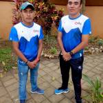 Two young men stand in a garden wearing blue and white Oax Sport shirts. The man on the left wears a blue cap with the Oax logo, a red watch, and jeans. The man on the right wears black sports pants with blue shoes. Both have their hands clasped in front. The background features colorful plants, trees, and a beige wall.