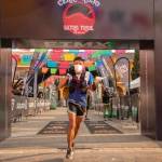 A runner wearing a blue shirt and black shorts crosses the finish line of the CECILIO Ultra Trail race. The runner, wearing a face mask and hydration pack, holds a map. The background features colorful banners and race sponsor logos, with other participants visible behind. The setting is vibrant and energetic, marking the end of a challenging race.