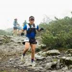 Corredor de Oax Sport Inc. participando en una carrera de trail en un entorno montañoso y neblinoso. El atleta lleva una camiseta azul y un chaleco de hidratación, con el número 270. En el fondo, se observan otros corredores en el recorrido.