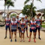 La imagen muestra a un grupo de atletas de Oax Sport Management posando juntos, todos vistiendo sus uniformes con los colores y logotipos del equipo. Están alineados en dos filas y todos llevan medallas alrededor del cuello, indicando que han completado una competencia. El fondo incluye palmeras y una estructura arquitectónica con arcos, lo que sugiere que están en un parque o una plaza pública. Los atletas parecen satisfechos y orgullosos de sus logros.