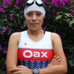 A female athlete from Oax Sport Management standing in front of vibrant purple flowers. She is wearing a white swim cap with goggles, and a white, red, and blue Oax Sport triathlon suit. Her arms are crossed, and she has a watch on her left wrist. She is looking directly at the camera with a slight smile.
