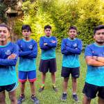 Five male athletes standing with arms crossed, wearing blue and teal jerseys with "Berrendo de Guía" logo. They pose confidently on a grassy lawn with a backdrop of lush green foliage and bamboo fencing. The group is aligned in a V-formation, showcasing unity and determination.