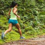 Mujer corriendo por un sendero verde en la naturaleza, vistiendo una camiseta sin mangas verde y shorts azules.