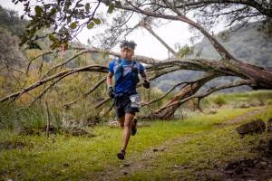 Corredor de trail running en un sendero boscoso, vistiendo camiseta azul y portando un chaleco de hidratación, rodeado de vegetación verde y árboles caídos en un entorno montañoso.