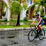 Oax Sport athlete cycling on a wet road, wearing a blue and white Oax Sport jersey and helmet, with palm trees and white archway buildings in the background.