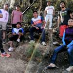 Group of athletes in Oax Sport gear posing outdoors in a forest area with relaxed expressions and camaraderie. The sun filters through trees, creating a rainbow effect. They are on a dirt path with trees and a rustic wooden structure in the background. The setting is natural and vibrant.