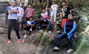 Group of athletes in Oax Sport gear posing outdoors in a forest area with relaxed expressions and camaraderie. The sun filters through trees, creating a rainbow effect. They are on a dirt path with trees and a rustic wooden structure in the background. The setting is natural and vibrant.