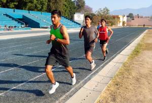 Three male runners in action on a track during a training session, with one wearing a green tank top, another in a gray shirt, and the third in a red shirt. The track is surrounded by bleachers, trees, and buildings, with mountains in the background.