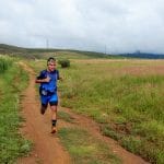 Male athlete running on a dirt path between cornfields and grassy fields, wearing a blue and black outfit, with hills and a cloudy sky in the background.