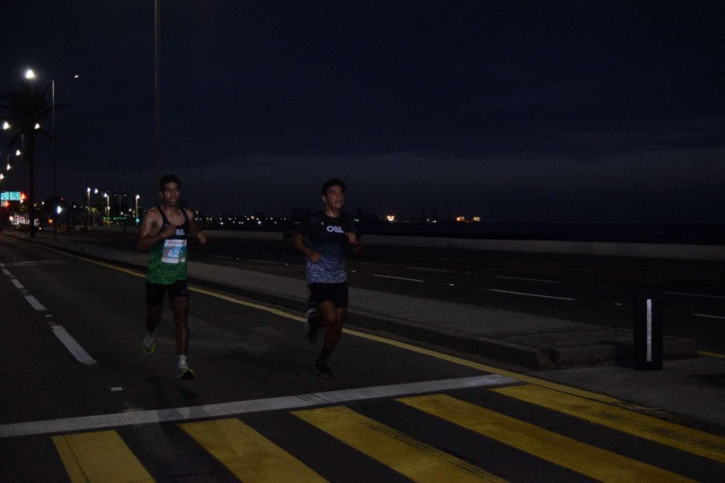 Two Oax Sport athletes running along the seafront during the Veracruz Marathon 2022 at night. The runner on the left wears bib number 622, and the road is illuminated by streetlights with a dark sky and distant city lights visible in the background.