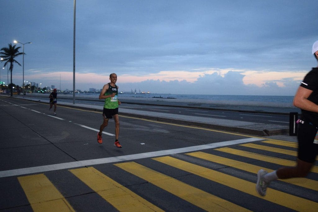 Oax Sport athlete running along the seafront during the Veracruz Marathon 2022, with bib number 945. The early morning sky is slightly illuminated, and the sea is visible in the background. The road is marked with yellow pedestrian lines.