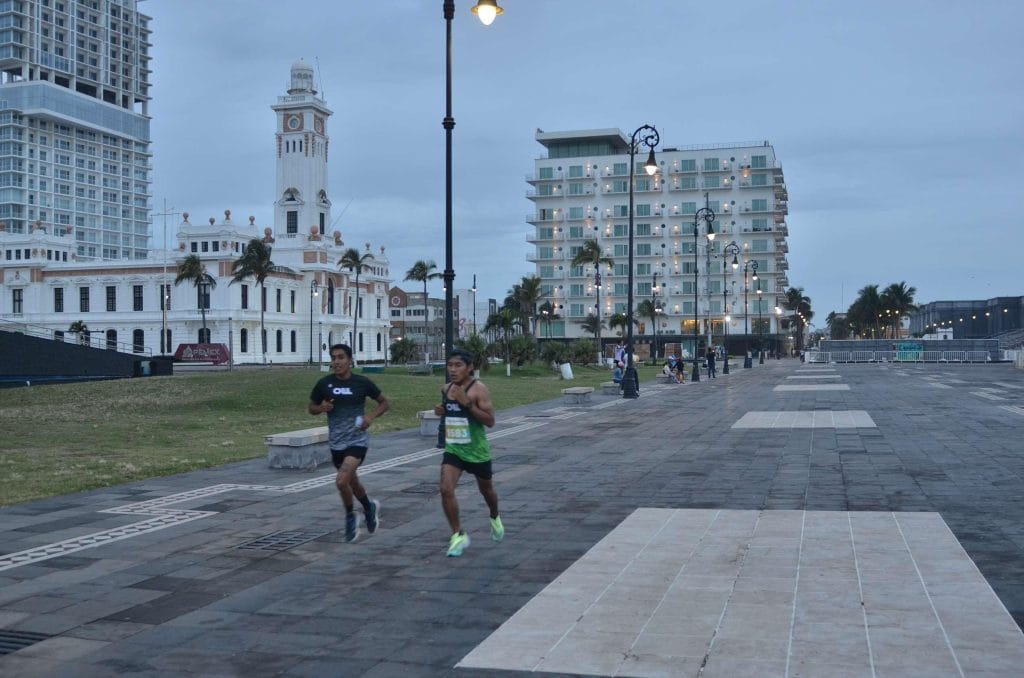 Two Oax Sport athletes running during the Veracruz Marathon 2022, with bib number 1583 leading. They are on a paved walkway with city buildings, including a clock tower, in the background. The sky is overcast, and streetlights are on.