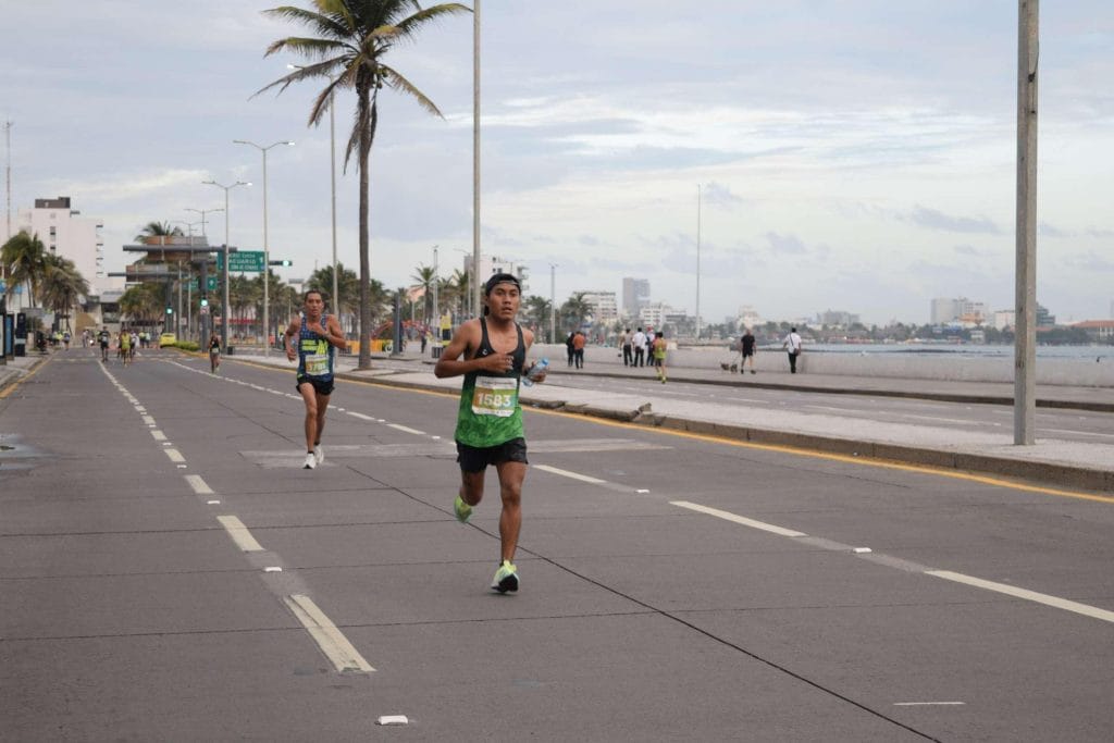 Runner with bib number 1583 participating in the Veracruz Marathon 2022, sprinting along a coastal road lined with palm trees and spectators. The sky is overcast, and the cityscape is visible in the background.