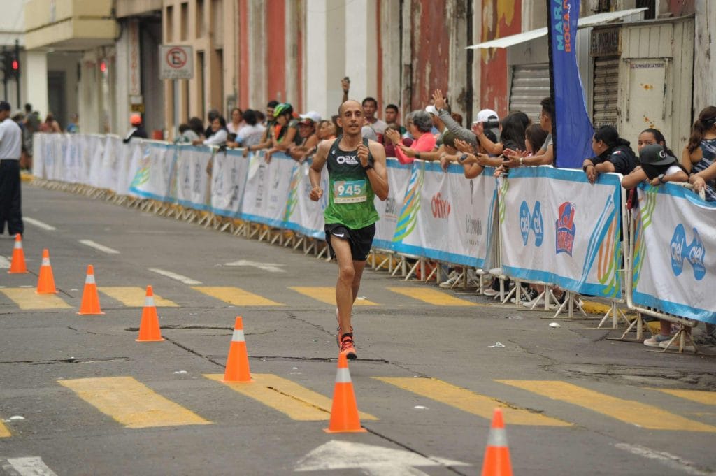 Runner nearing the finish line at the Veracruz Marathon 2022, cheered on by enthusiastic spectators along the route. The runner is wearing a green Oax Sport shirt and is focused on completing the race. Orange cones line the path, guiding the runners to the finish.