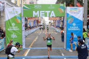Runner crossing the finish line at the Veracruz Marathon 2022, under the "META" banner. Other participants are seen in the background with spectators cheering from the sidelines. The event is sponsored by Casas Bambino and Veracruz. The runner wears a green Oax Sport shirt and appears elated with his performance.