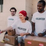 Three volunteers standing at a table with aid supplies, including boxes labeled "Medicine" and "Fragile." The woman in the middle, wearing a red bandana, is handing a bottle of water to someone off-camera. The man on the left has glasses, and the man on the right has a beard. All three are wearing white shirts with "VOLUNTEER" printed on them. They are in an outdoor setting next to a building.