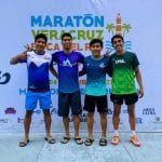 Four Oax Sport athletes stand in front of a Maratón Veracruz Boca del Río banner, smiling and making hand gestures. They wear different athletic shirts and shorts, showing camaraderie and readiness for the marathon.