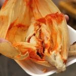 Una persona abriendo un tamal de salsa roja en un plato de plástico blanco.