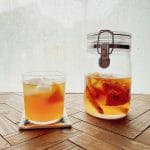 A glass jar with a metal clasp and a glass filled with a cold drink containing chunks of fruit, both placed on a wooden surface with a light background.