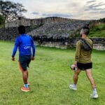 Dos corredores, uno con camiseta azul de Oax Sport y otro con camiseta verde, caminando en un campo de césped junto a antiguas ruinas de piedra bajo un cielo parcialmente nublado.