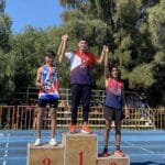 Three athletes standing on a podium, celebrating their victories. The first-place winner is in the center, wearing a red and purple uniform with a gold medal. The second-place athlete, on the left, is wearing a red, white, and blue uniform with a silver medal. The third-place athlete, on the right, is wearing a red and blue uniform with a bronze medal. Trees and a blue sky are in the background.