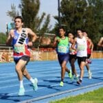 Juan Zurita Victoria, in a green and blue uniform, competing in a track race alongside other athletes on a blue track. He is running with determination, surrounded by fellow competitors, under a clear sky with trees in the background.