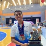 Juan Zurita Victoria, an athlete from Magdalena Apasco, Etla, Oaxaca, proudly holding his trophies and medals in an indoor sports arena. He is wearing a white and blue La Salle University jersey, with a big smile on his face.