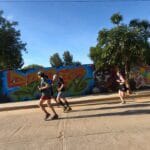 Charlie in green, Ollie Bishop from England in black, Luis Fabian Santiago in blue, and Anaya from England in black running past a colorful mural during the Monte Alban Sunrise Run in Oaxaca.