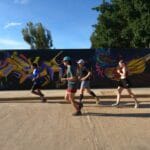 Luis Fabian Santiago in a blue shirt, Charlie from Georgia in green, Ollie Bishop from England in black, and Anaya from England in black running past a vibrant mural during the Monte Alban Sunrise Run in Oaxaca.