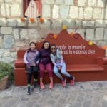 Three women sitting on a red bench against a stone wall, with a sign behind them that reads "diablo la muerte mientras la vida nos dure." From left to right: Mane Peña, Diana Garcia, and Lisa from Scouts Design.