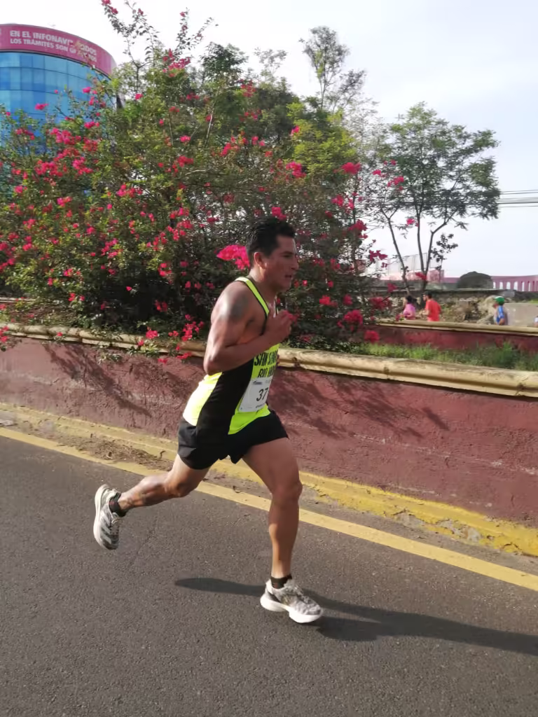 Noel Cirenio Fabian Hernandez running in a race, showcasing his determination and athleticism against a backdrop of vibrant pink flowers and a cityscape in San Sebastián Río Hondo.