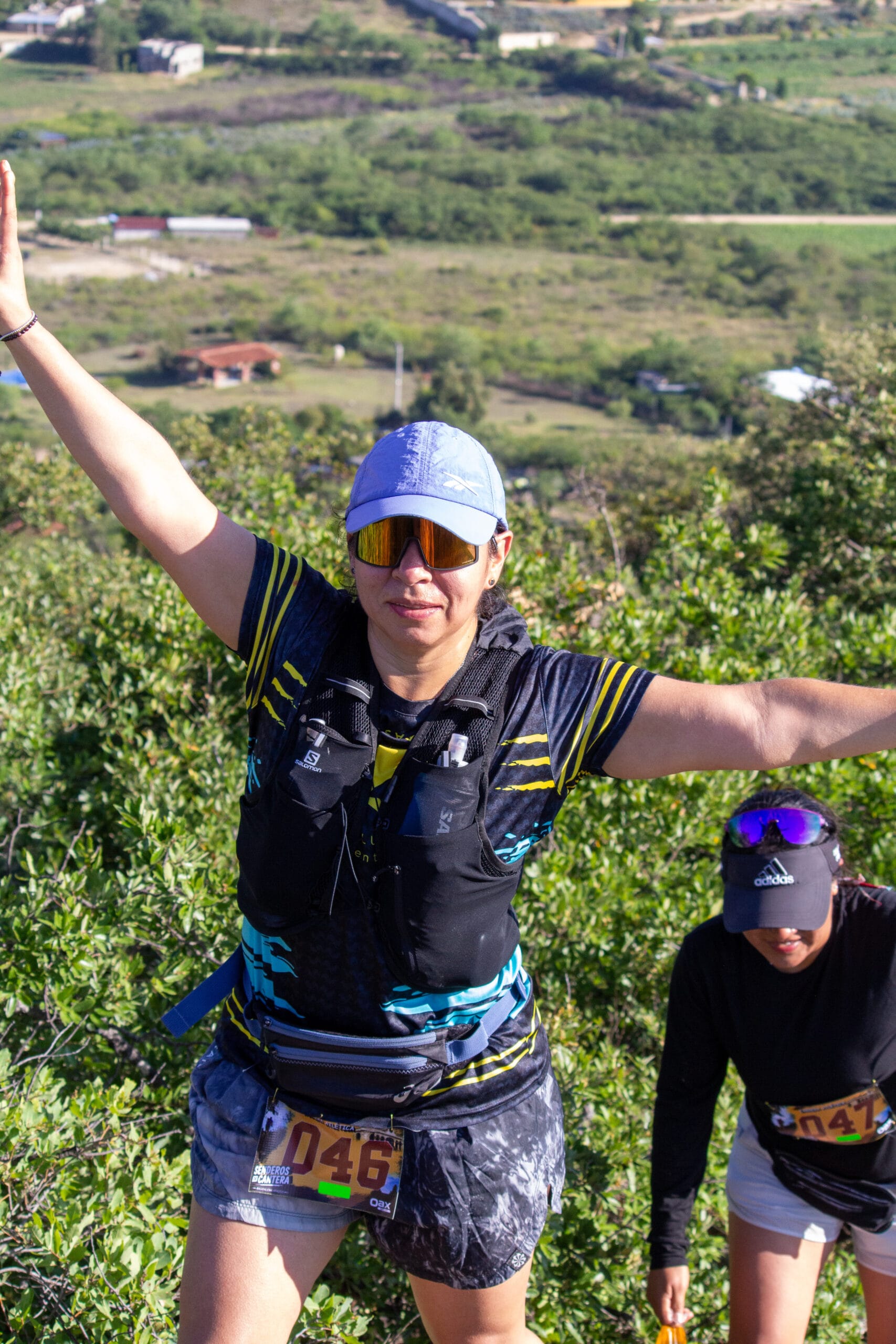 Dos corredores suben por un sendero empinado sobre los campos de Magdalena Apazco; el corredor que va adelante levanta ambos brazos.