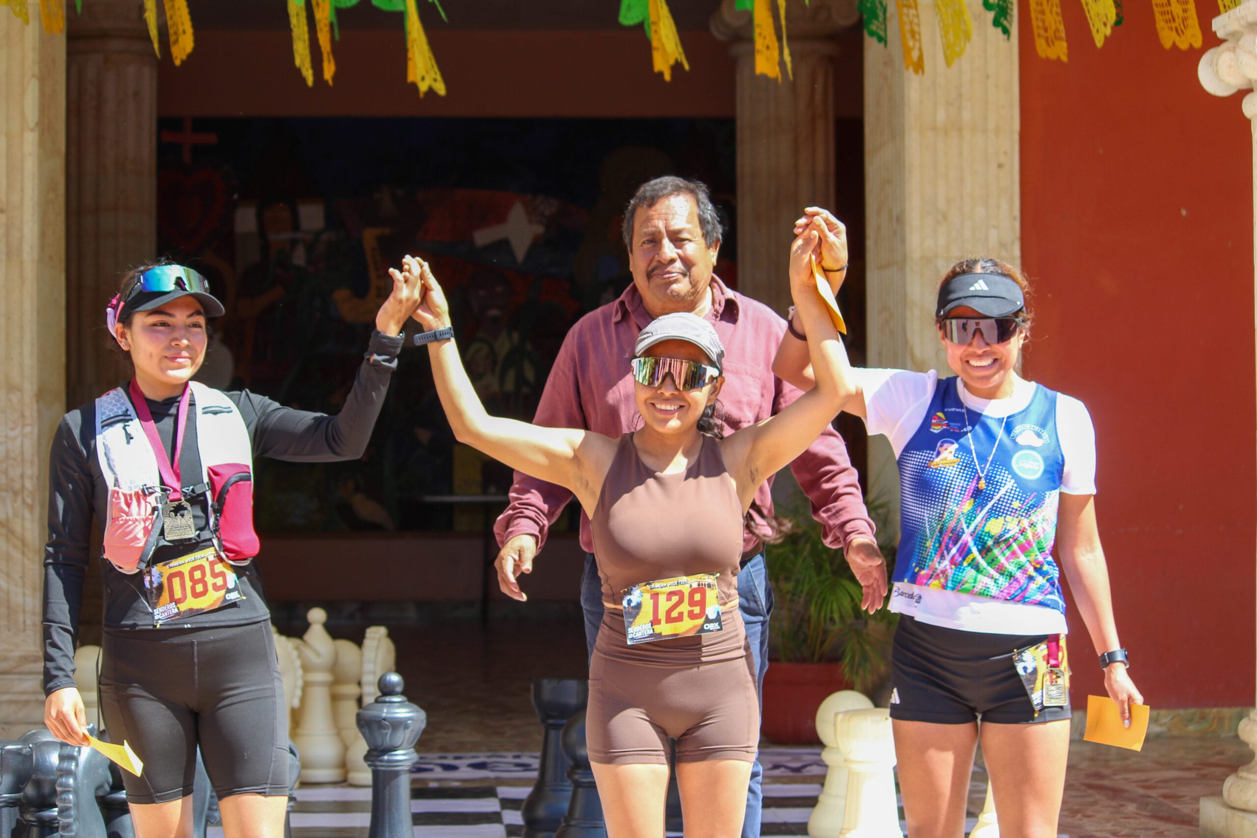 Tres corredoras con medallas celebran durante la premiación mientras una autoridad municipal aparece detrás de ellas.