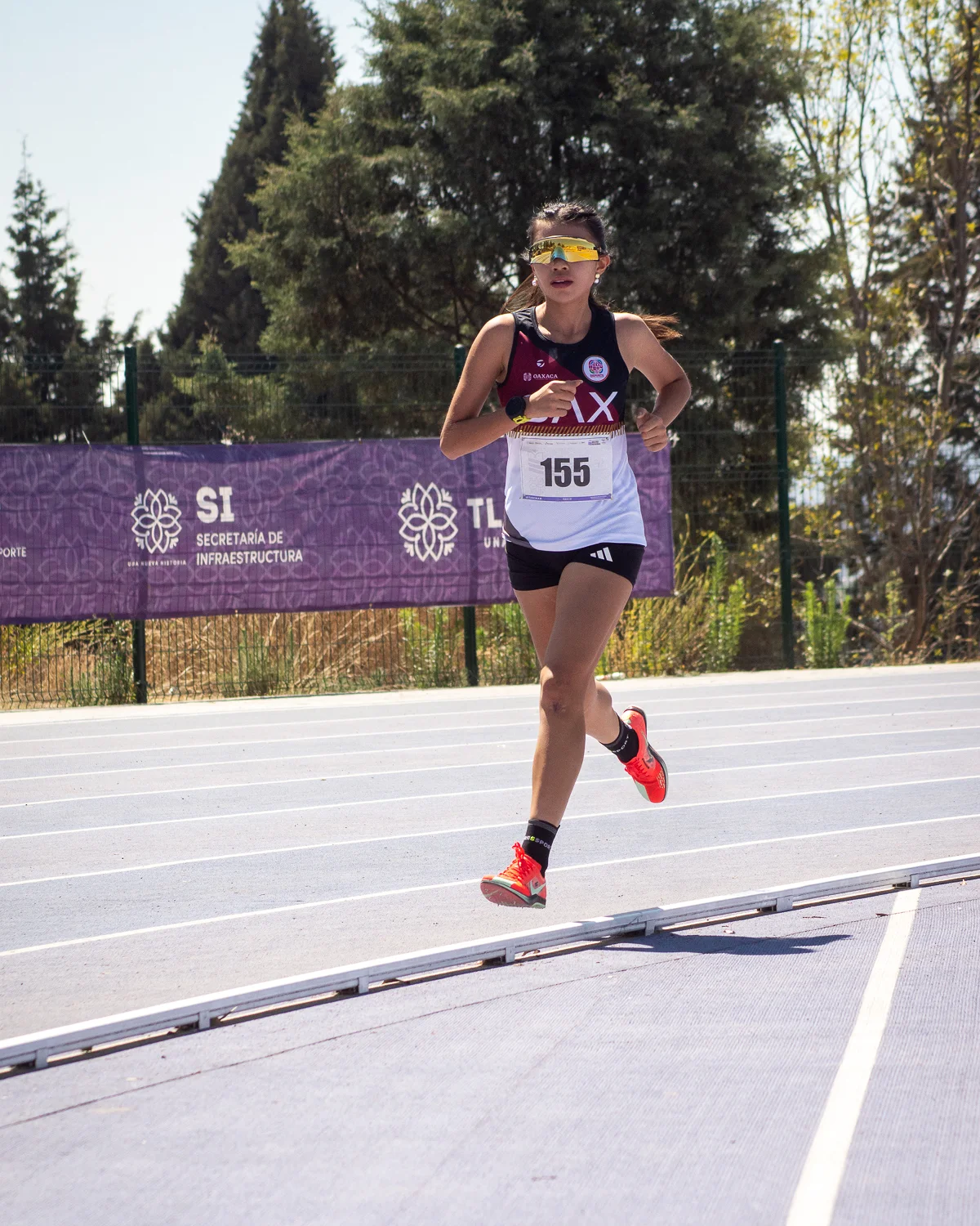 Carolina Cinco running on a track during a race, wearing bib 155 and yellow sunglasses.
