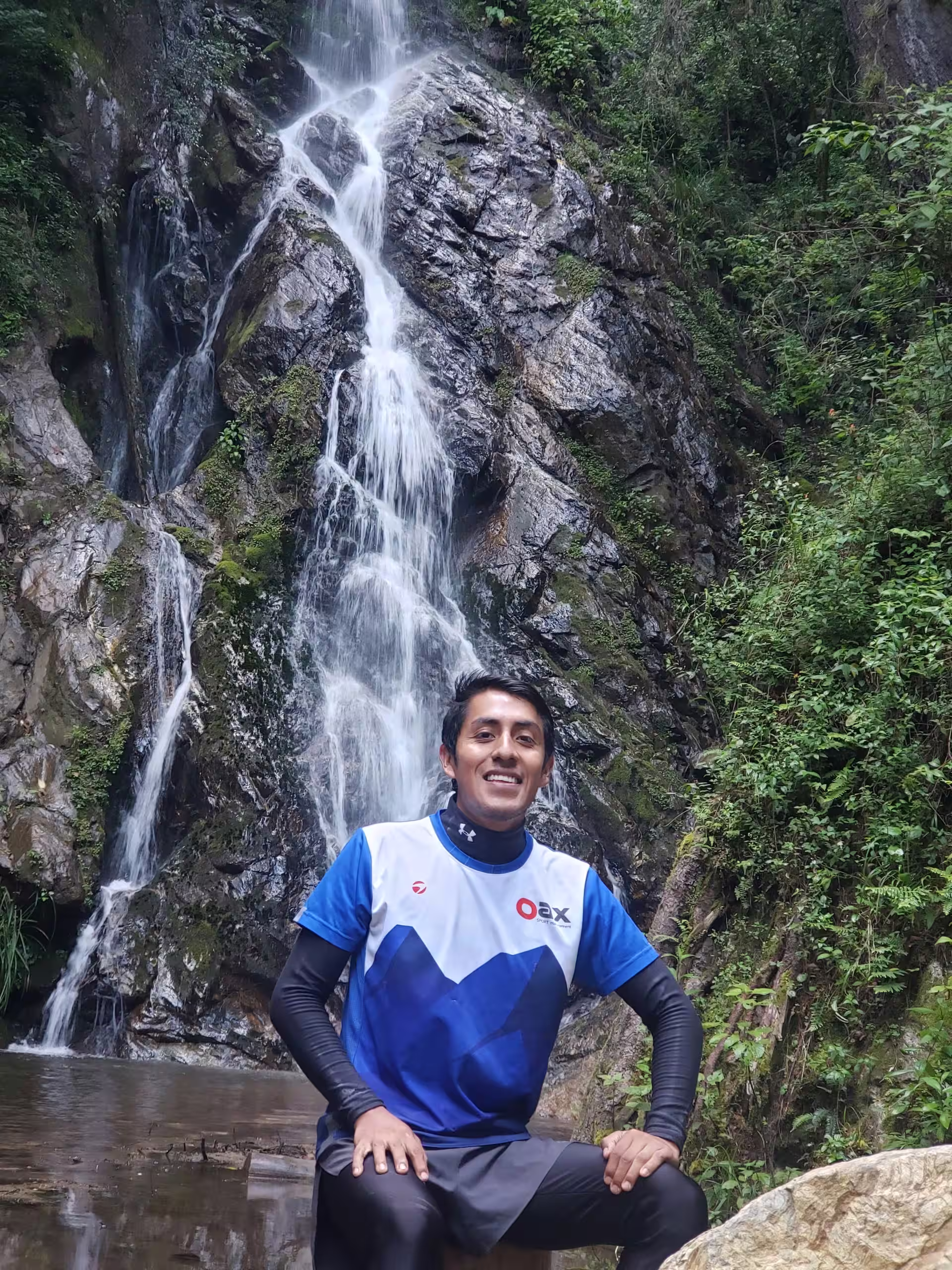 Un corredor con playera azul de Oax Sport, sentado frente a una cascada en la montaña.